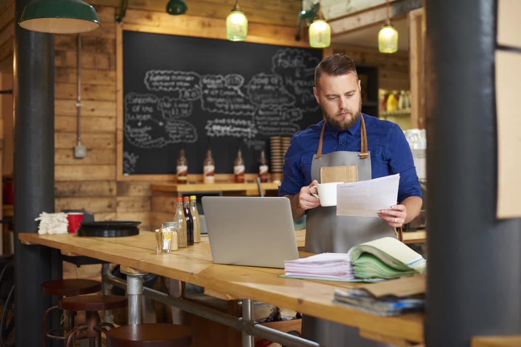 a coffee shop owner checks the delivery notes in his bookkeeping folder on the counter of his busy coffee shop and cross checks them with his online accountancy via his laptop at the end of a busy day .