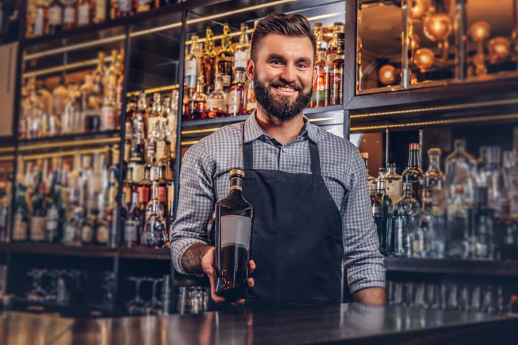 Cheerful bartender in apron presents a bottle of exclusive alcohol at bar counter background.